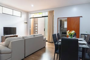 A modern living room with a sectional sofa, a dining table with chairs, a flat-screen TV, and a sliding glass door leading to a kitchen area.