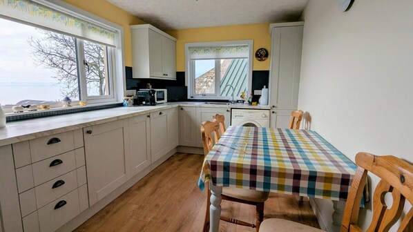 Une cuisine avec des meubles blancs, un four à micro-ondes, un évier et une table recouverte d’une nappe à carreaux.