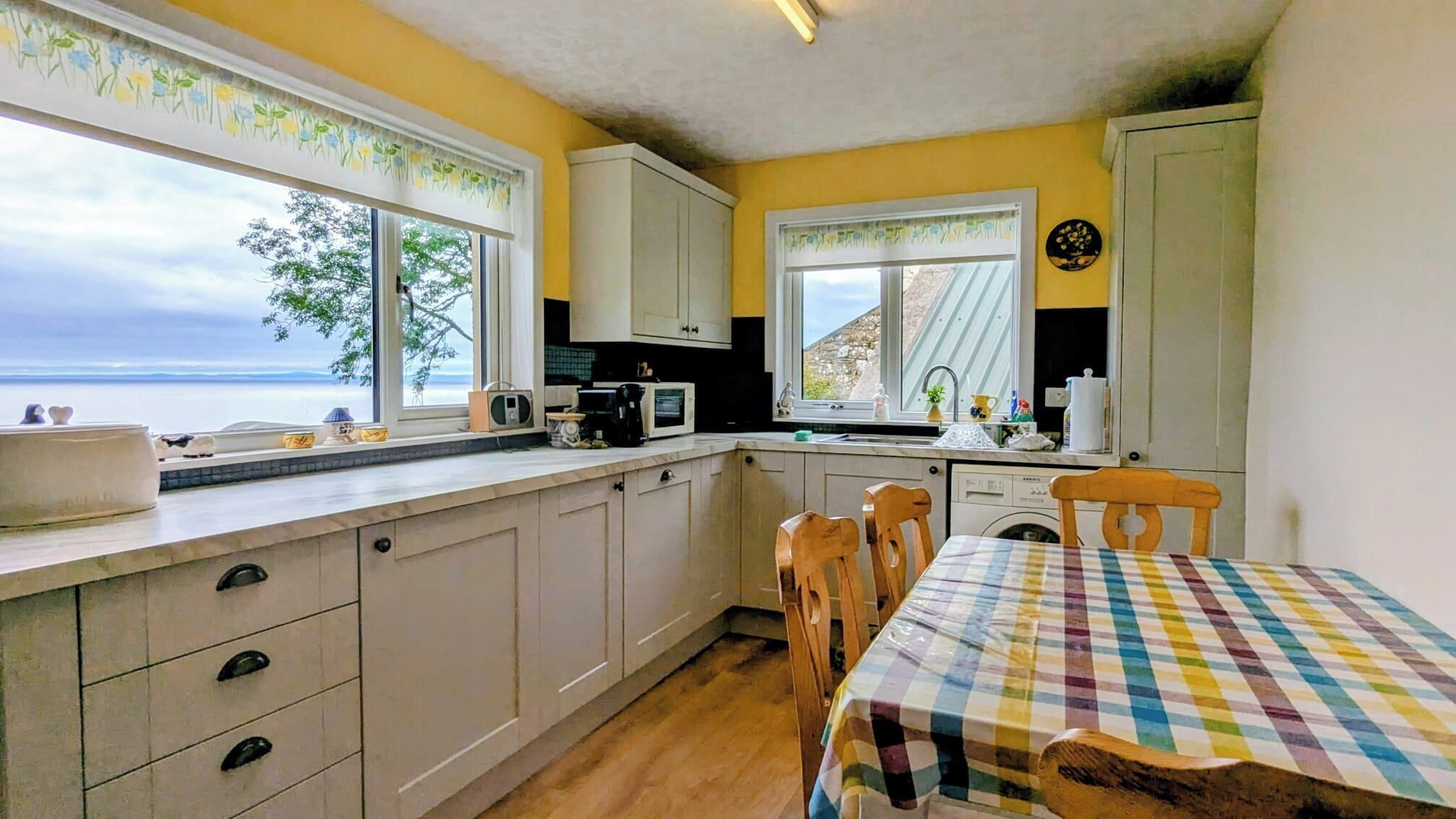 A kitchen with a view of the sea, a dining table with a checkered tablecloth, and wooden chairs.