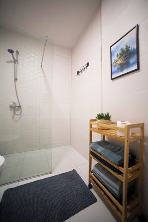 A modern bathroom with a glass shower enclosure, a wooden shelf with towels, a framed landscape painting, and a toilet. - Modern Family Suite in Prizren (Prizren)