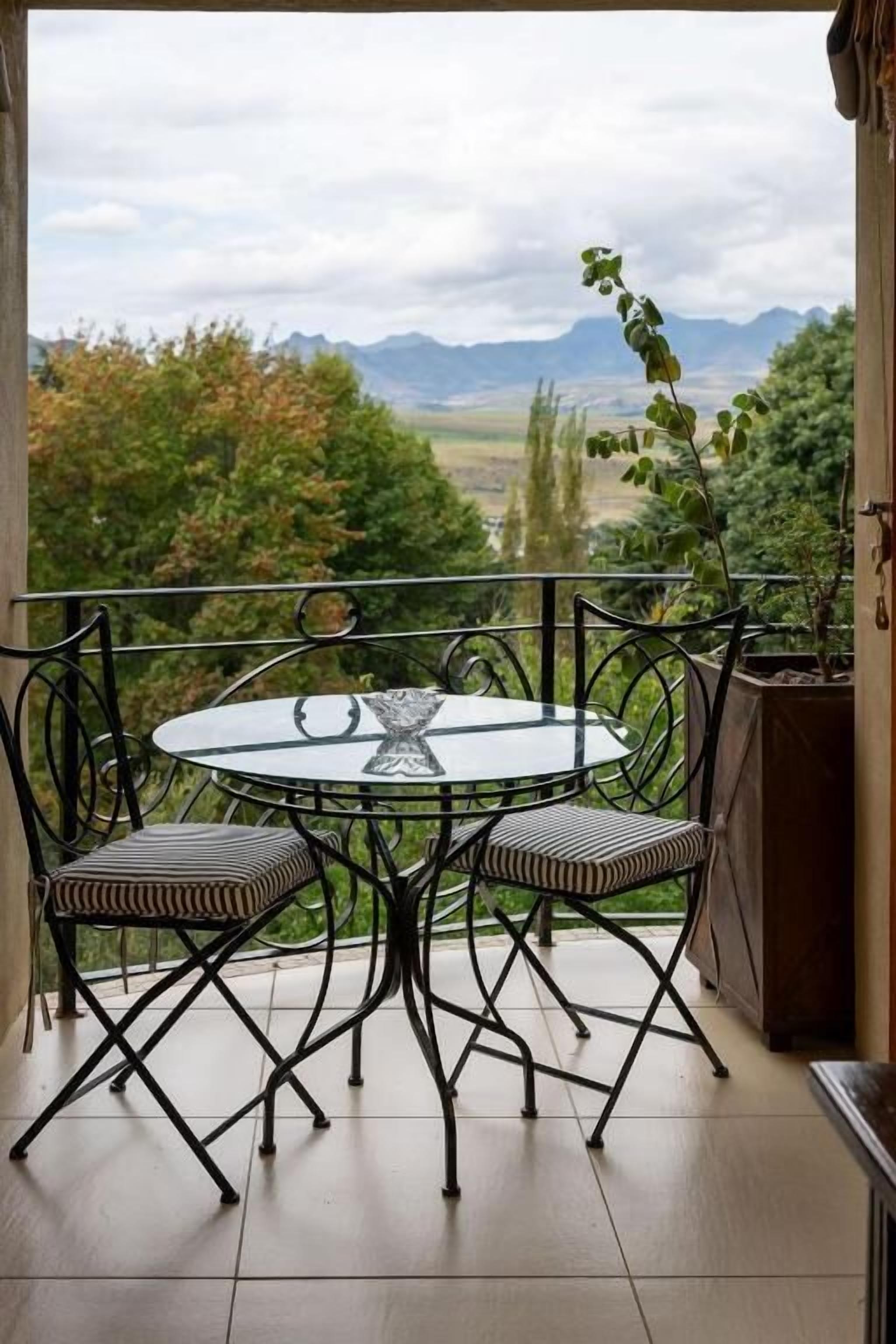 Un balcon avec une table ronde et des chaises, un pot de fleurs et une vue sur les montagnes et les arbres.