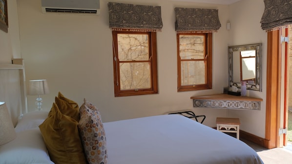 A bedroom with a bed, two windows with curtains, a bedside table, a mirror, and an air conditioning unit.