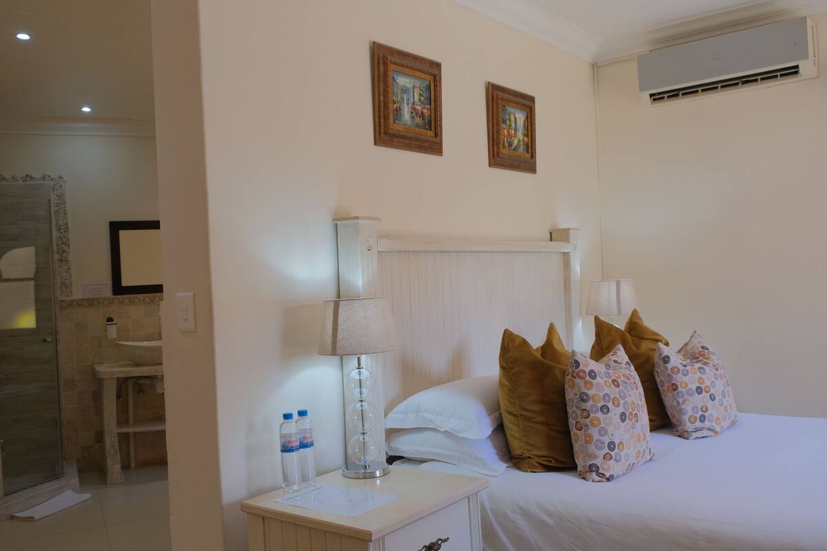 A hotel room with a bed, pillows, a nightstand, a lamp, a mirror, and a shower area.