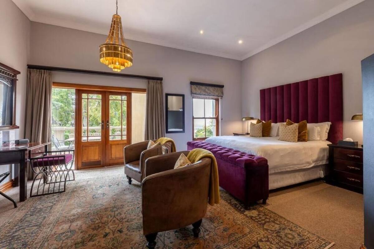 A bedroom with a bed, a desk, two armchairs, a chandelier, and a view of the outdoors.