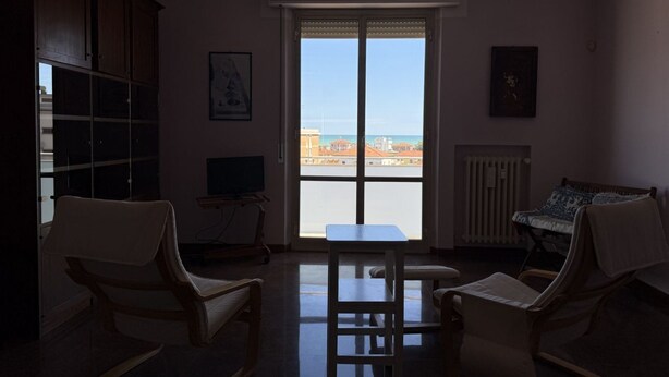 A room with a view through a large window, a table, two chairs, a television, and a radiator. - Location: in a residential area, a few steps from the sea and all services.Description: apartment on the fourth floor of a building with lift. Living room with terrace overlooking the sea - bright kitchen with dining table and balcony - double bedroom wi (SENIGALLIA)