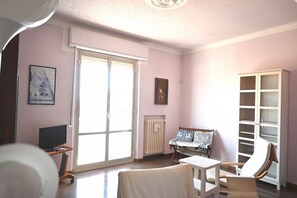 Een kamer met een televisie, een stoel, een tafel, een radiator en een groot raam.