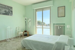 Een kamer met een bed, een stoel, een radiator, een raam met uitzicht en een lijst met een afbeelding aan de muur.