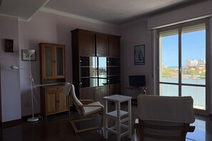 Une pièce avec une grande fenêtre, un meuble, une télévision et deux chaises.