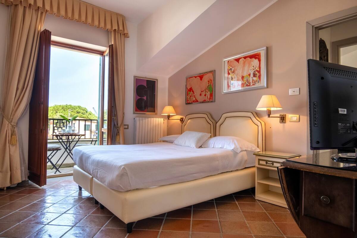 A hotel room with a bed, two bedside lamps, a TV, a small table, and a balcony with a chair.