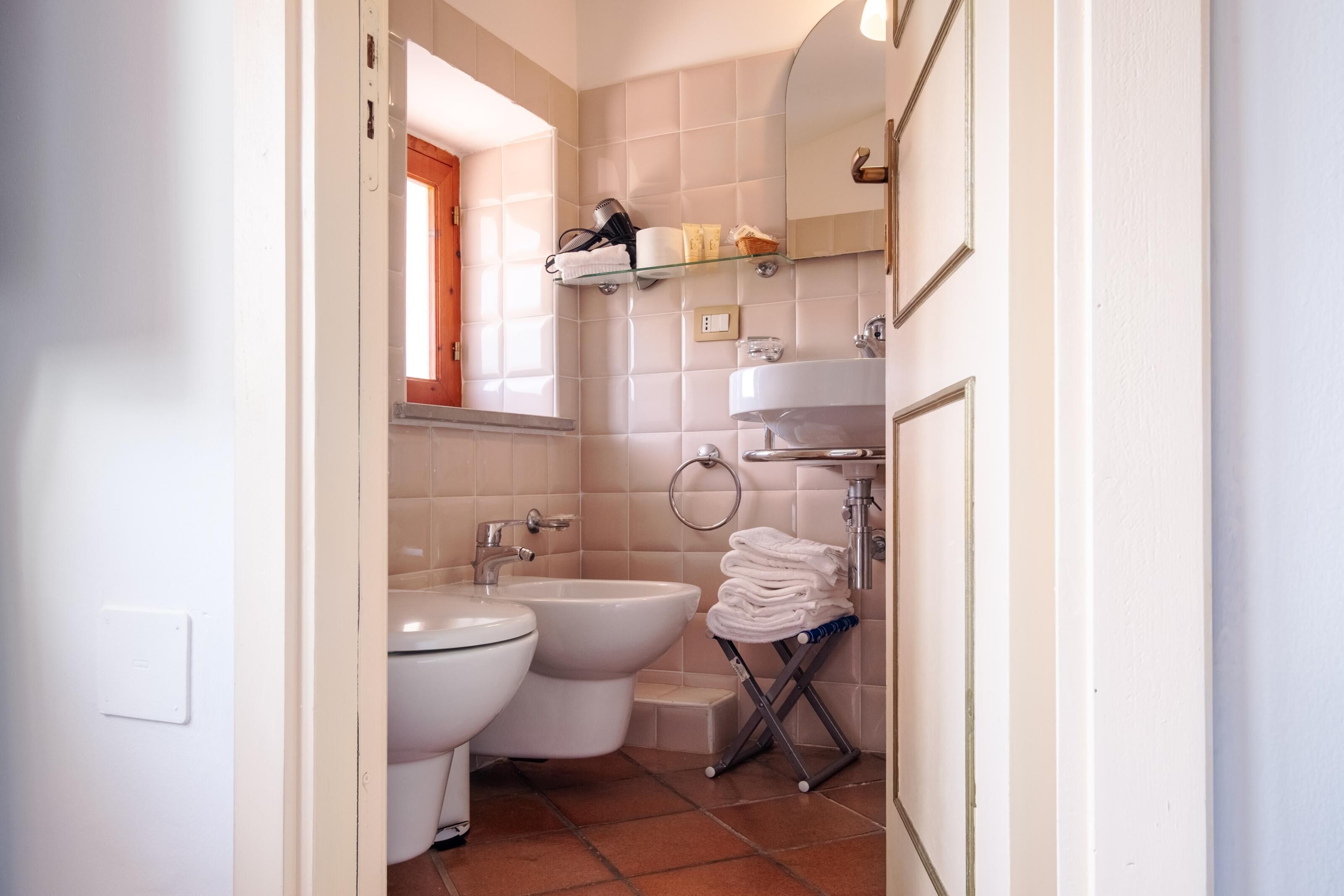 A bathroom with a toilet, bidet, and sink. There is a towel rack with towels and a shelf with toiletries.
