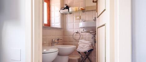 A bathroom with a toilet, bidet, and sink. There is a towel rack with towels and a shelf with toiletries.