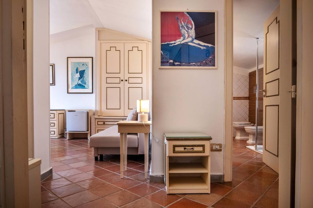 A hallway with a bed, a bedside table, a lamp, a framed picture, and a door leading to a bathroom.