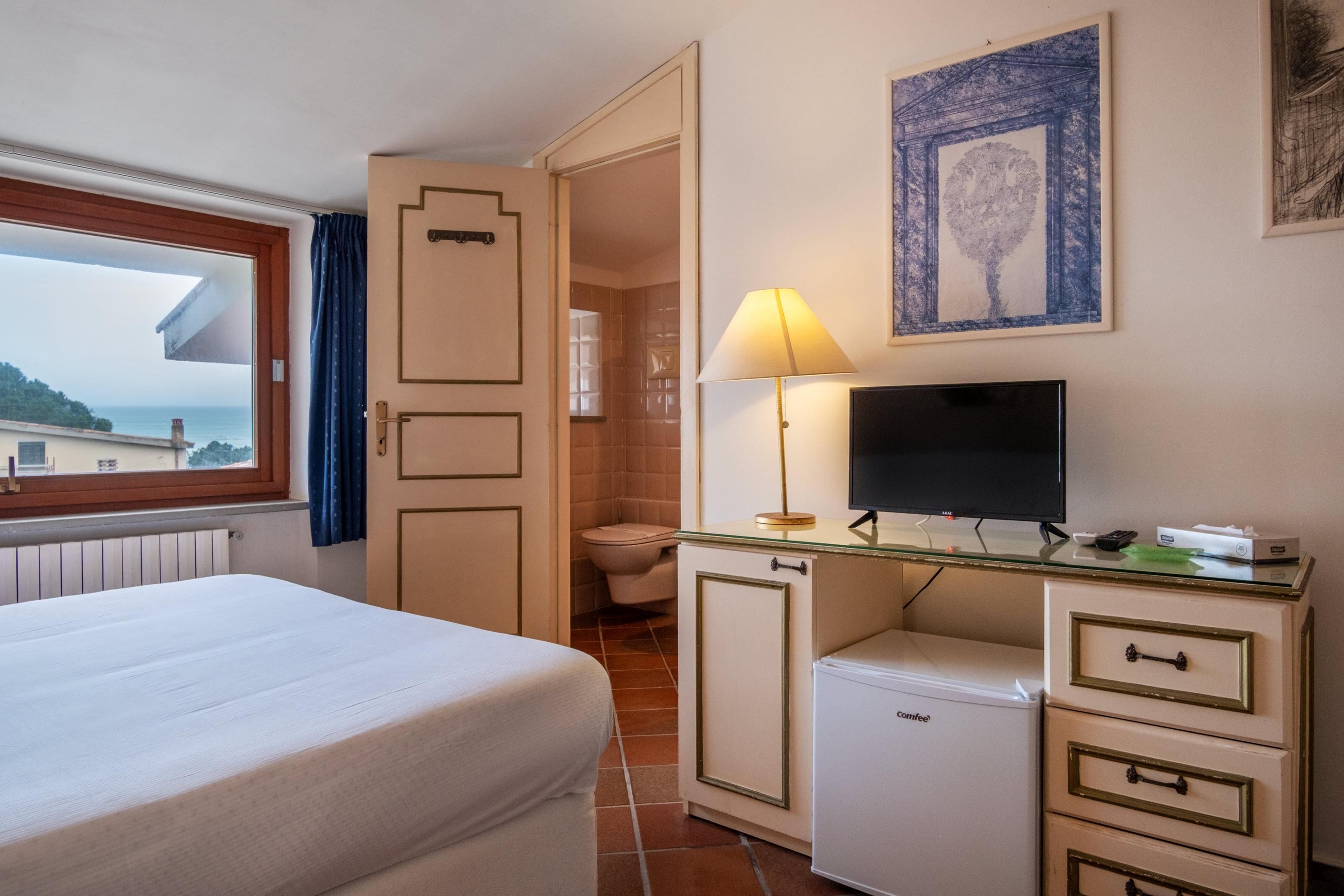 A hotel room with a bed, a desk with a television, a small refrigerator, and a bathroom visible through an open door.