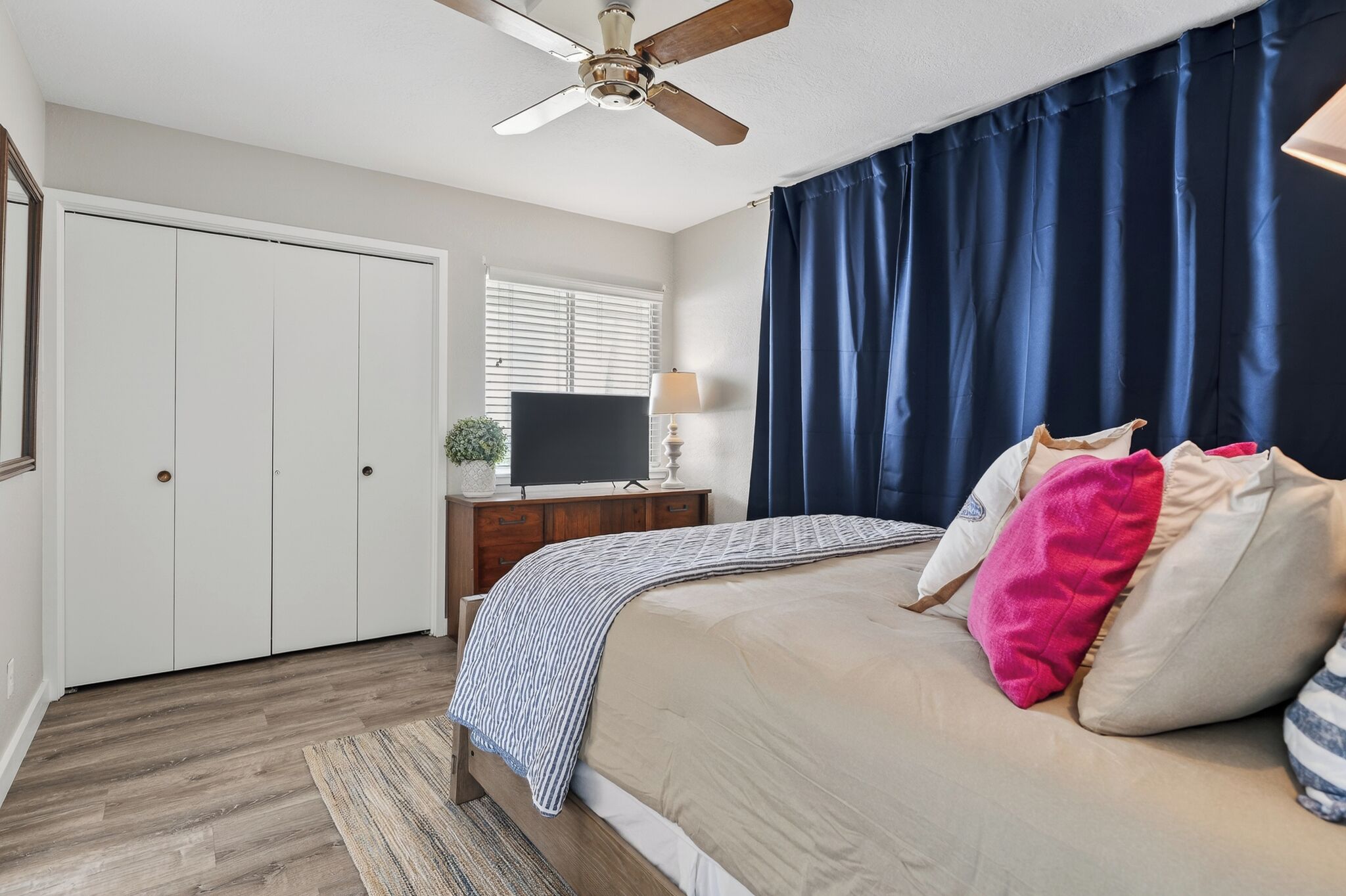 Un dormitorio con cama, ventilador de techo, televisor sobre un mueble de madera, ventana con persianas y una puerta.