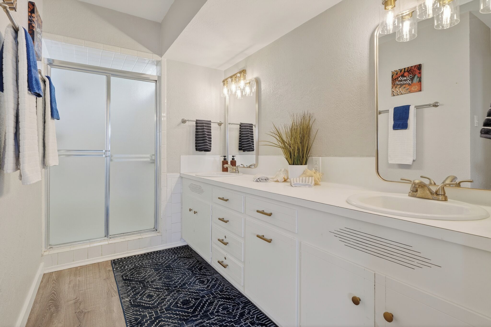 A modern bathroom with a glass shower door, white vanity with gold handles, and a large mirror with lights.