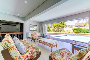 Uma sala de estar moderna com vista para a piscina, área de bar e mobiliário com temática tropical.
