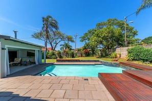 A swimming pool with a wooden deck, a covered patio with chairs, and a view of a residential area with trees and a clear sky.