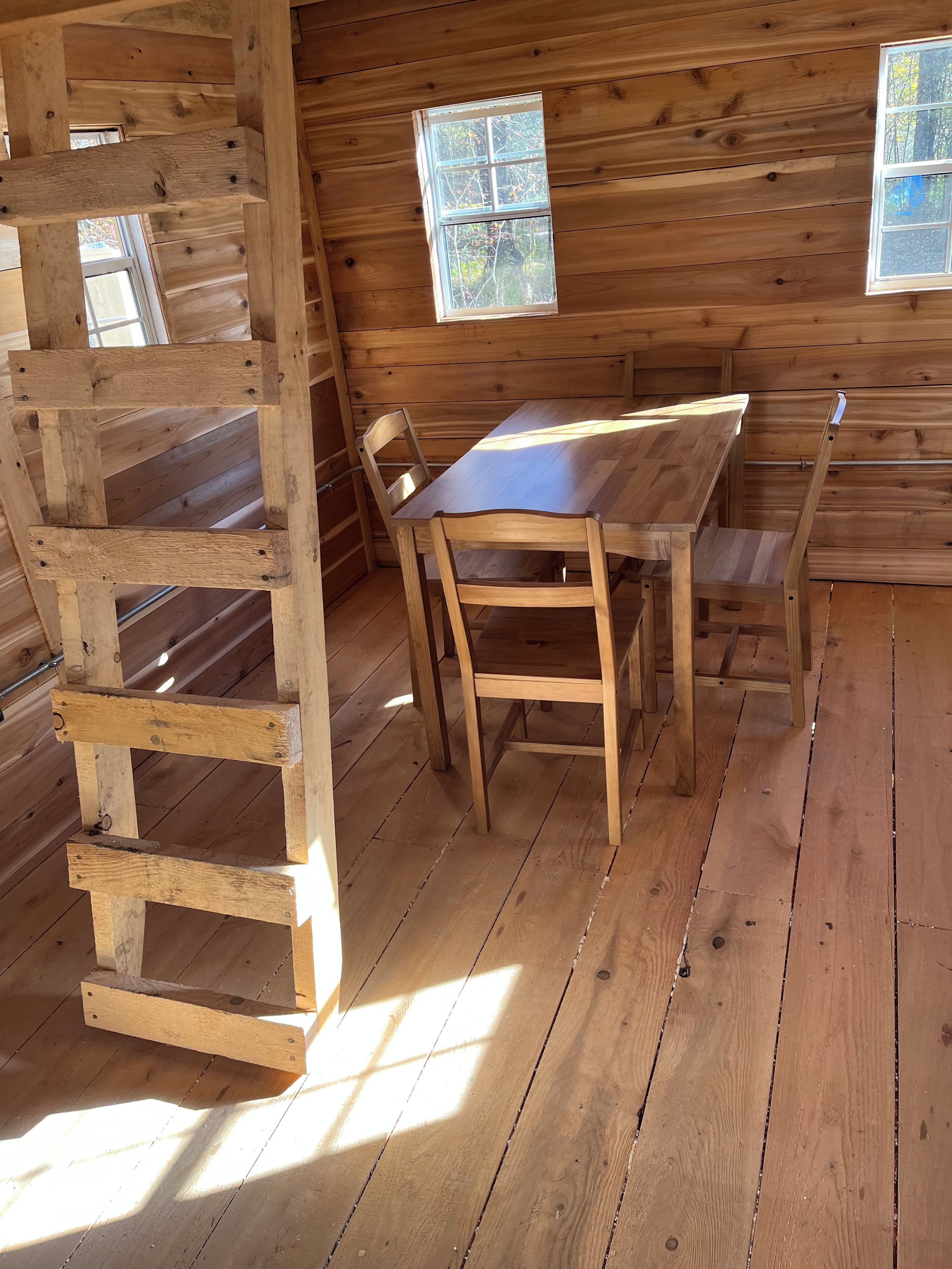 A wooden interior with a table and chairs, a wooden ladder, and two windows.