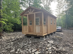 A wooden cabin with a porch, windows, and a door, surrounded by trees and gravel.