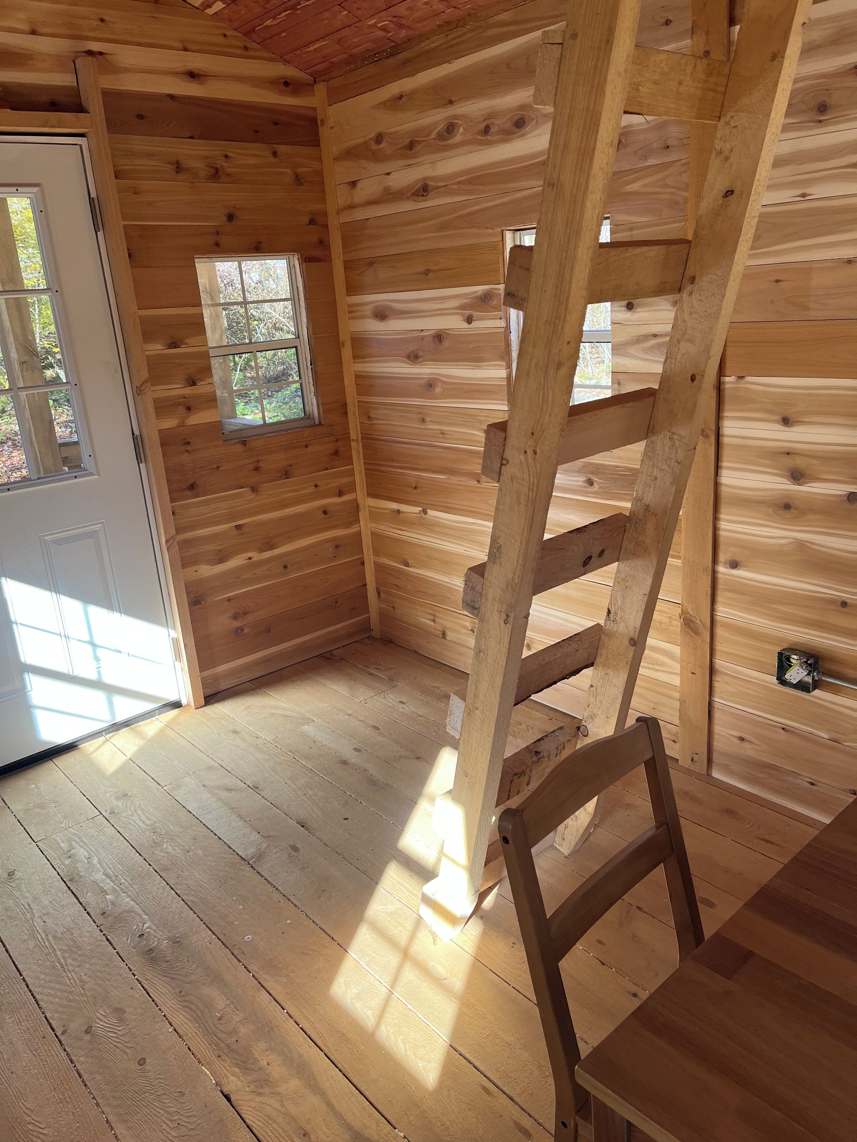 A wooden cabin interior with a wooden ladder, a door, and a window.