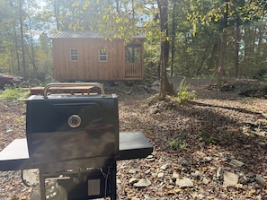 A portable grill in a wooded area with a wooden cabin in the background.