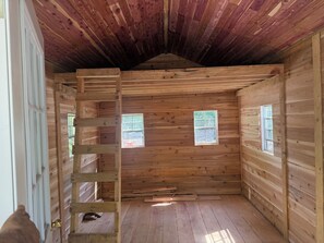 A wooden cabin interior with a loft area, two windows, and a door.