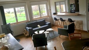 A modern living room with a sofa, chairs, a dining table, and large windows offering a view of the outdoors.