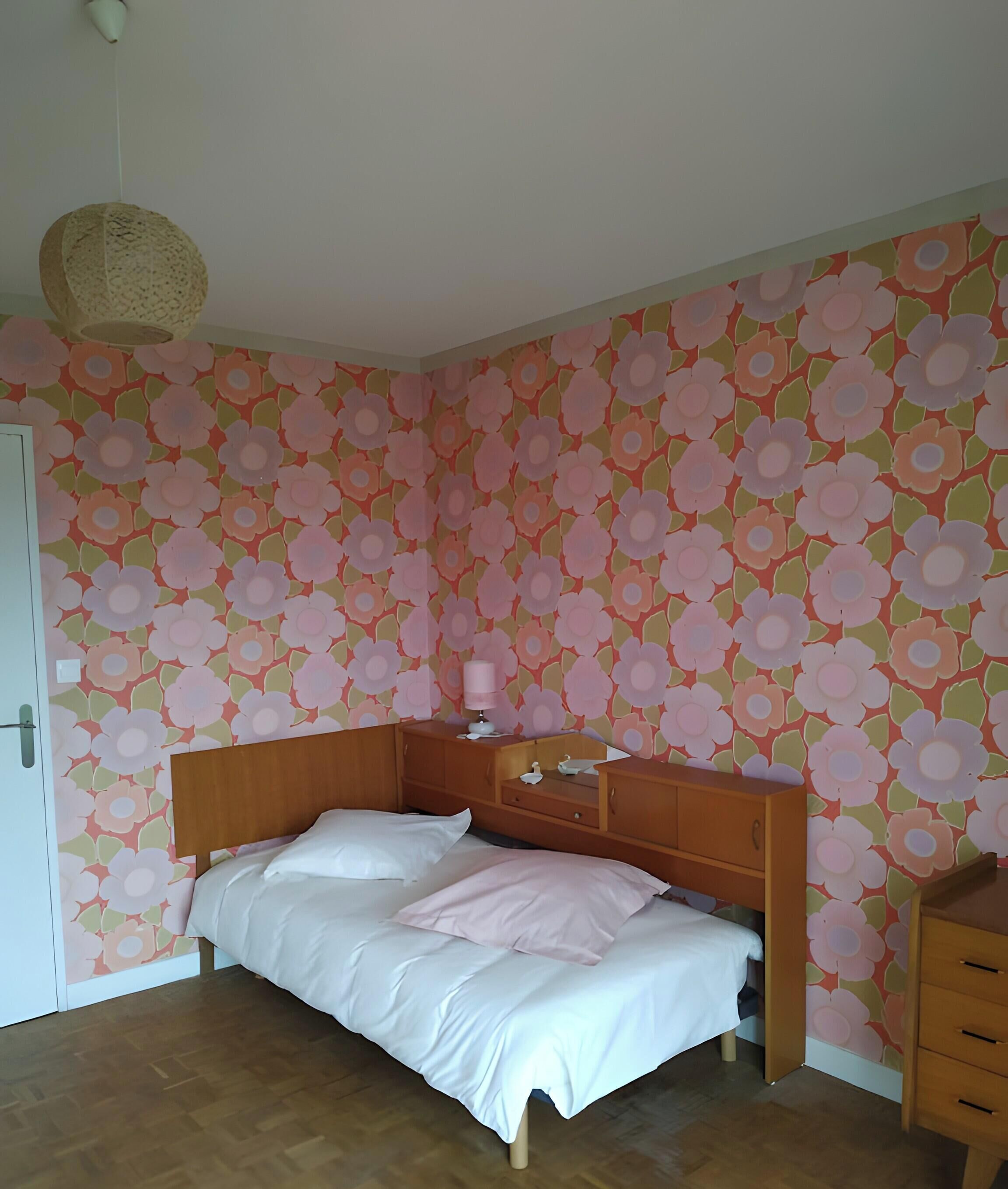 A bedroom with a wooden bed, a bedside table, a lamp, and a floral wallpaper.