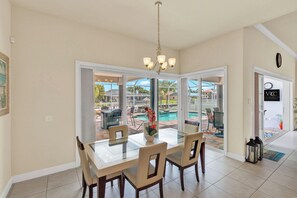 Un espace repas avec une table et des chaises en verre, un lustre, et une vue sur une piscine et un coin salon extérieur.