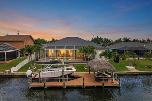A waterfront property with a swimming pool, a boat docked at the pier, and a thatched-roof structure.