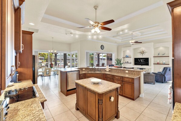 A spacious kitchen with granite countertops, wooden cabinets, and a central island. There is a dining area with chairs and a large window.