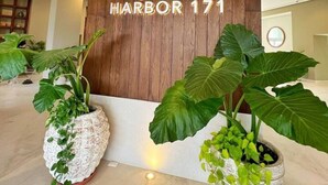 Een receptie met een houten wand en een groot verlicht 'M'-logo, potplanten en een bord waarop 'HARBOR 171' staat.