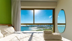 Ett hotellrum med ett stort fönster, en säng med vita sängkläder, utsikt över stadsbilden och en spegel på väggen.