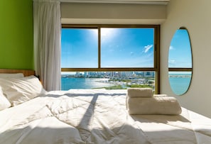 A hotel room with a large window, a bed with white linens, a view of a cityscape, and a mirror on the wall. - Parque do Cais #1109 With Sea View by Carpediem (Recife)