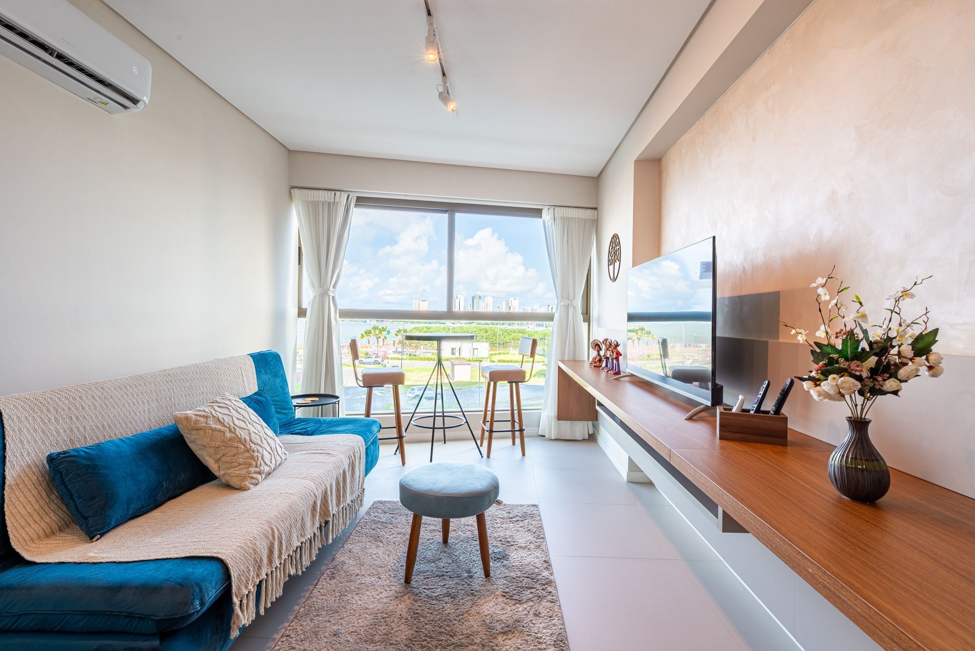Una sala de estar moderna con un sofá, reposapiés, televisor y vista de la ciudad a través de amplios ventanales.
