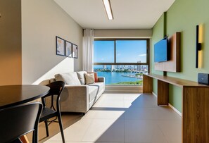 A modern living room with a sofa, a dining table, a TV mounted on the wall, and a view of a marina. - Parque do Cais #1109 With Sea View by Carpediem (Recife)