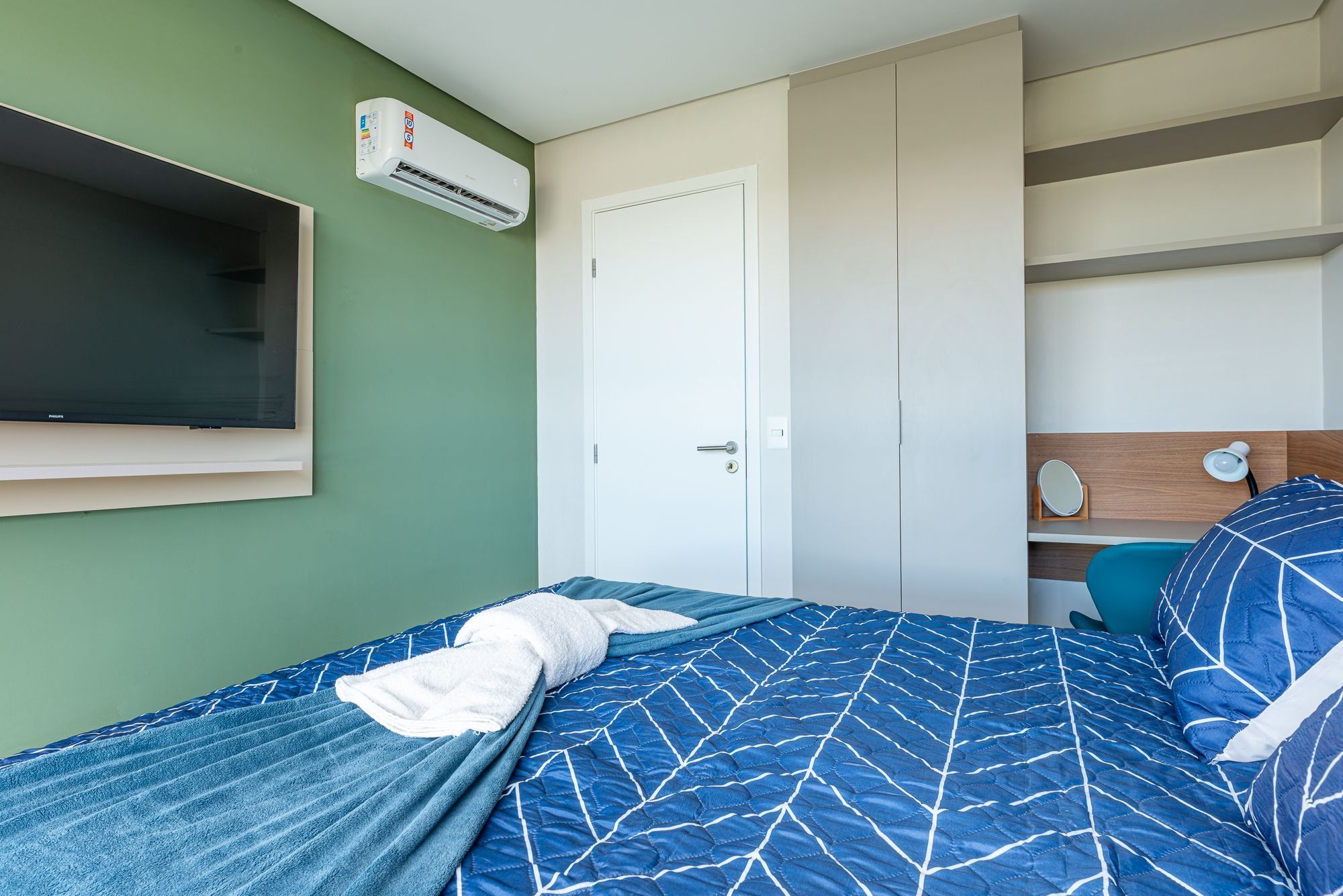 Un dormitorio con cama, televisor, aire acondicionado, puerta y mesita de noche con lámpara.