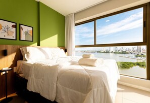 Quarto de hotel com uma cama grande, paredes verdes, obras de arte e vista para a cidade e para a água.