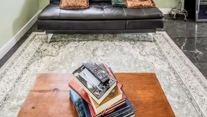 Una sala de estar con un sofá de cuero negro, una mesa de centro con libros, un tapete estampado y vistas al exterior con vegetación.