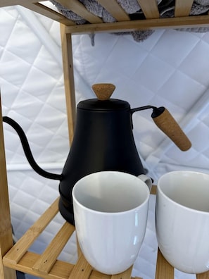 A black coffee maker with a cork handle on a wooden shelf, two white cups, and a white quilted wall in the background.