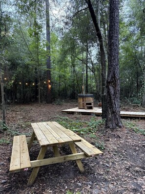 A forested area with a wooden picnic table and a small wooden structure, possibly a fireplace or heater, surrounded by trees.