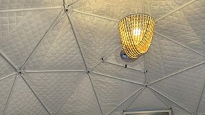 Um quarto com tenda, teto acolchoado, porta com vidro jateado, luminária pendente, prateleira com objetos e uma cama branca.
