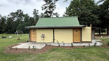 A small wooden building with a green roof, a concrete pathway leading to it, and a grassy area with trees in the background.