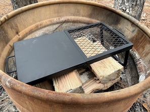A metal grill with a mesh top, placed over a fire pit with logs.