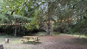 A picnic table in a forested area with string lights overhead. - Canvas Cabin with Camp Grill and Cozy Outdoor Retreat in Branchville, South Carolina (Branchville)
