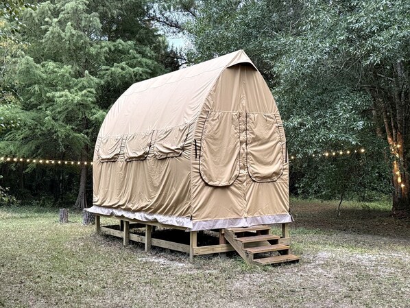 A covered wooden structure with a flat roof, supported by wooden legs, surrounded by trees and string lights.