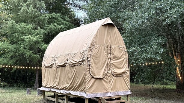 A covered wooden structure with a flat roof, supported by wooden legs, surrounded by trees and string lights. - Canvas Cabin with Camp Grill and Cozy Outdoor Retreat in Branchville, South Carolina (Branchville)