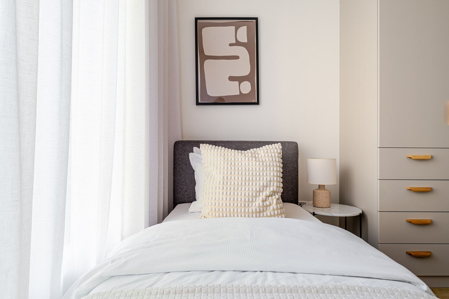 Una cama bien tendida con un almohadón, una mesita de noche con lámpara y un armario con tiradores de madera.