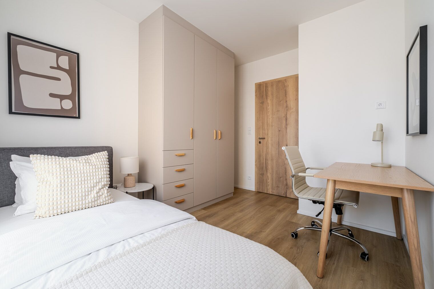 Ein Schlafzimmer mit Bett, Nachttisch, Lampe, Kleiderschrank, Schreibtisch, Stuhl und einer Tür.