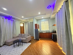 Uma sala de estar com sofá, mesa de centro e uma área de cozinha com geladeira, micro-ondas e pia.